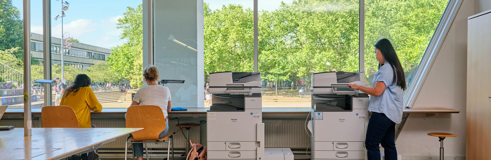 Eine Frau bedient einen Kopierer in einem hellen Raum mit großen Fenstern, zwei weitere Personen sitzen an einem Tisch und arbeiten