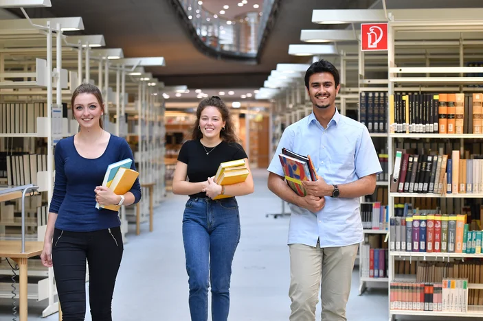 Drei Studierende mit Büchern in der Hand laufen nebeneinander im Gang in der Zentralbiblitothek der SUB Göttingen