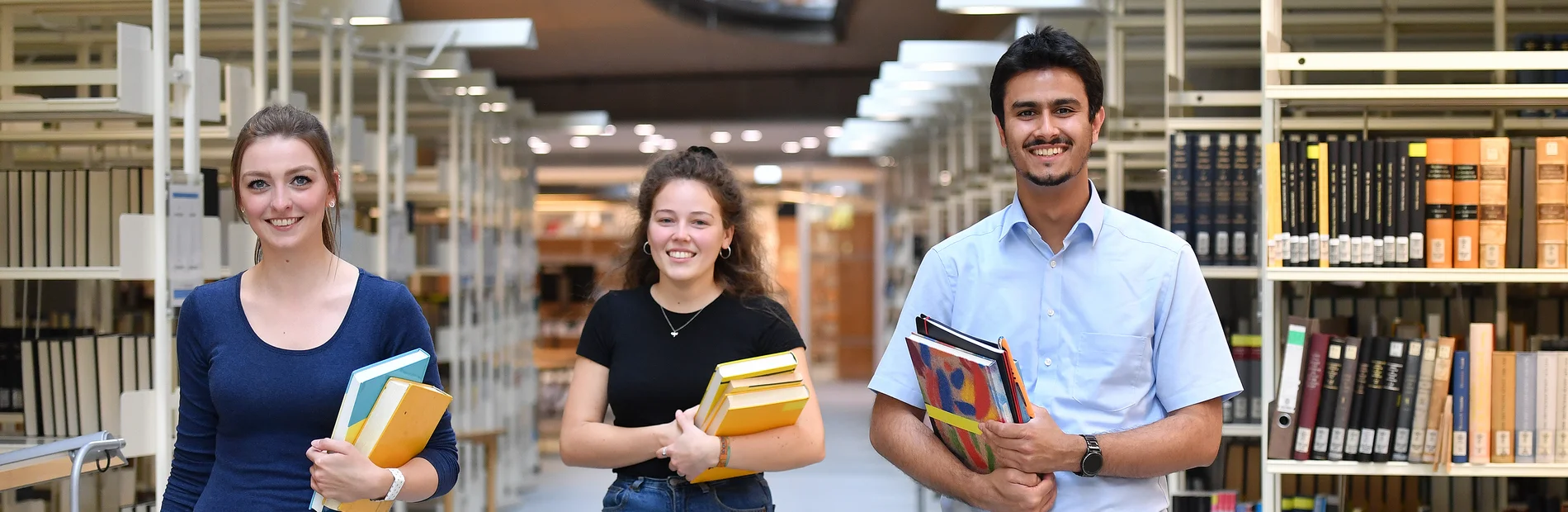 Drei Studierende mit Büchern in der Hand laufen nebeneinander im Gang in der Zentralbiblitothek der SUB Göttingen
