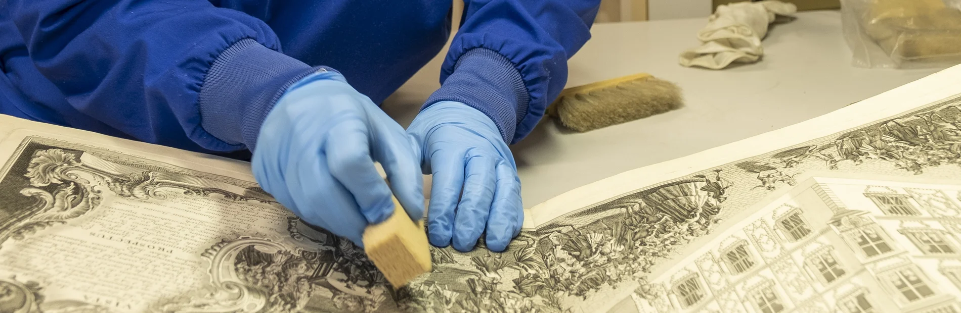 Trockenreinigung Zwei Hände in blauen Handschuhen reinigen mit einem Kautschukschwamm einen Kupferstich mit Gebäudedarstellung.