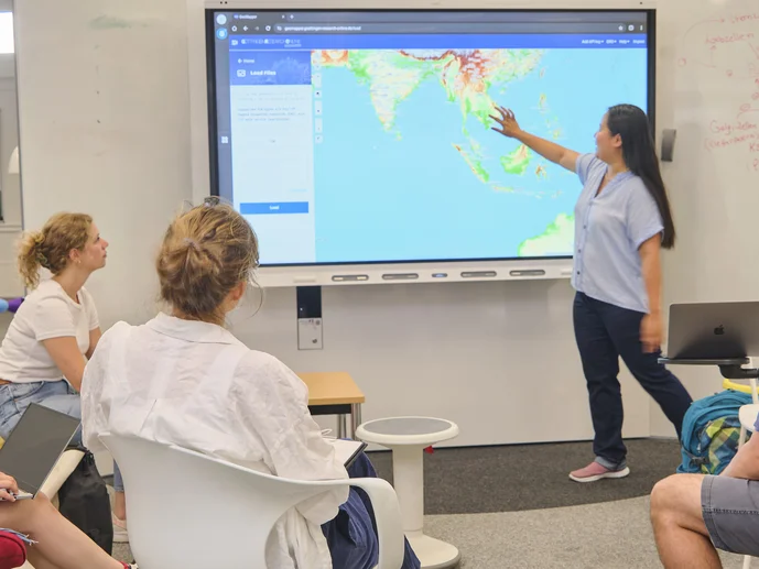 Frau präsentiert Weltkarte auf großem Bildschirm im Digital Creative Space, vier Personen sitzen und hören zu