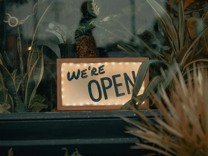 Beleuchtetes Schilf mit der Aufschrift "We're open", das in einem mit Pflanzen dekorierten Fenster steht.