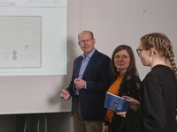 Vor einer Powerpointpräsentation stehen auf jeder Seite drei Menschen, die die Powerpoint betrachten. Diese zeigt eine historische Urkunde.