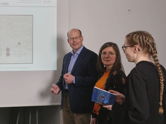 Kurse zur Arbeit mit historischen Beständen Vor einer Powerpointpräsentation stehen auf jeder Seite drei Menschen, die die Powerpoint betrachten. Diese zeigt eine historische Urkunde.