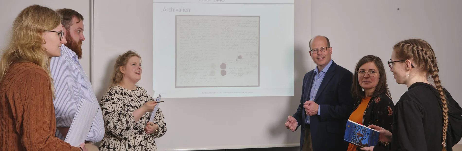 Kurse zur Arbeit mit historischen Beständen Vor einer Powerpointpräsentation stehen auf jeder Seite drei Menschen, die die Powerpoint betrachten. Diese zeigt eine historische Urkunde.