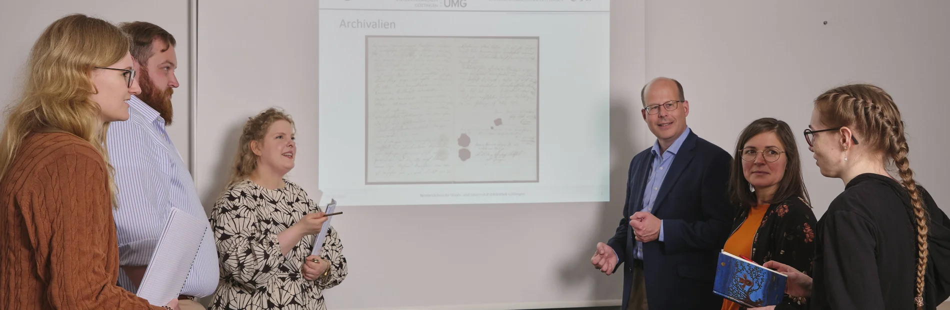 Vor einer Powerpointpräsentation stehen auf jeder Seite drei Menschen, die die Powerpoint betrachten. Diese zeigt eine historische Urkunde.