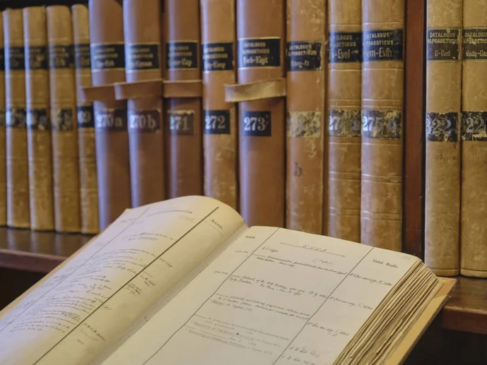 Im Hintergrund Bibliothekskataloge auf einem Regal, im linken vorderen Bereich ein aufgeschlagener Bibliothekskatalog