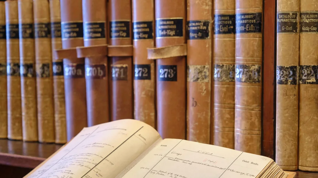 Im Hintergrund Bibliothekskataloge auf einem Regal, im linken vorderen Bereich ein aufgeschlagener Bibliothekskatalog