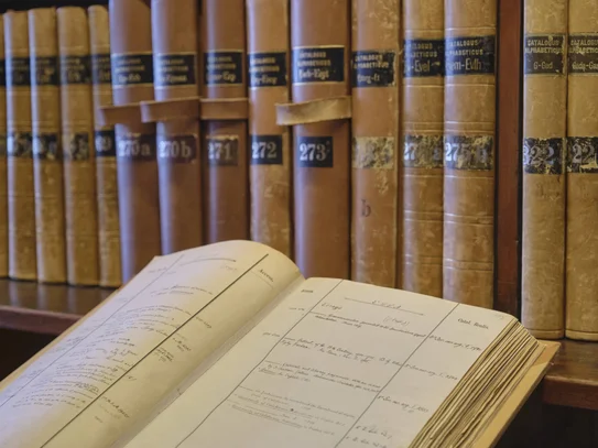 Recherchieren Im Hintergrund Bibliothekskataloge auf einem Regal, im linken vorderen Bereich ein aufgeschlagener Bibliothekskatalog
