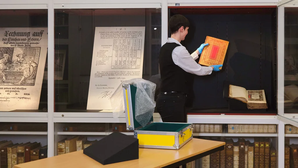 Restauratorin legt ein Buch in eine offene Vitrine; daneben geschlossene Vitrinen mit Faksimiles.