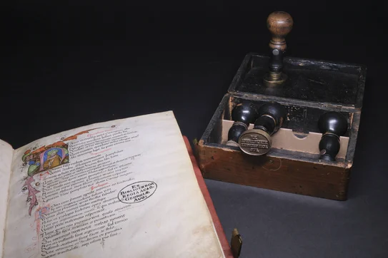 Links eine aufgeschlagene Handschrift mit dem Stempel „EX BIBLIOTHECA REGIA ACAD. REGIAE AUG:“, rechts ein Holzkasten mit historischen Stempelwerkzeugen.