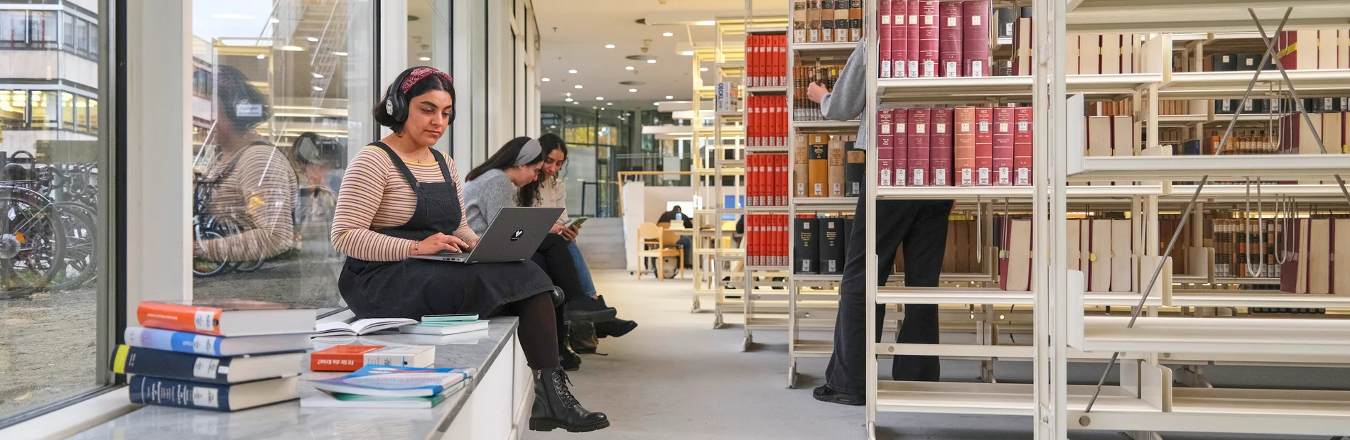 Zwei Frauen sitzen in der Bibliothek auf einer Fensterbank, eine arbeitet an einem Laptop, im Hintergrund Bücherregale und weitere Personen