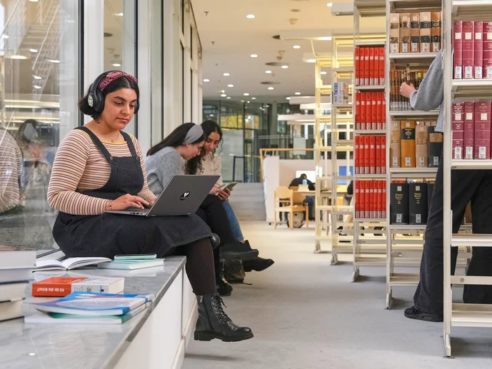 Zwei Frauen sitzen in der Bibliothek auf einer Fensterbank, eine arbeitet an einem Laptop, im Hintergrund Bücherregale und weitere Personen