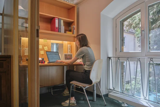 Arbeitskabine im HG der SUB Göttingen Das Bild zeigt das Innere einer Arbeitskabine. Eine Studentin sitzt an einem Tisch und benutzt einen Laptop. Auf dem Tisch sind verschiedene Arbeitsmaterialien verteilt. Auf dem Regal über dem Tisch stehen mehrere Ordner und Bücher.