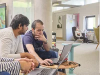 Drei Personen sitzen im Digital Creative Space an einem Tisch, zwei nutzen Laptops, eine Person zeigt auf den Bildschirm