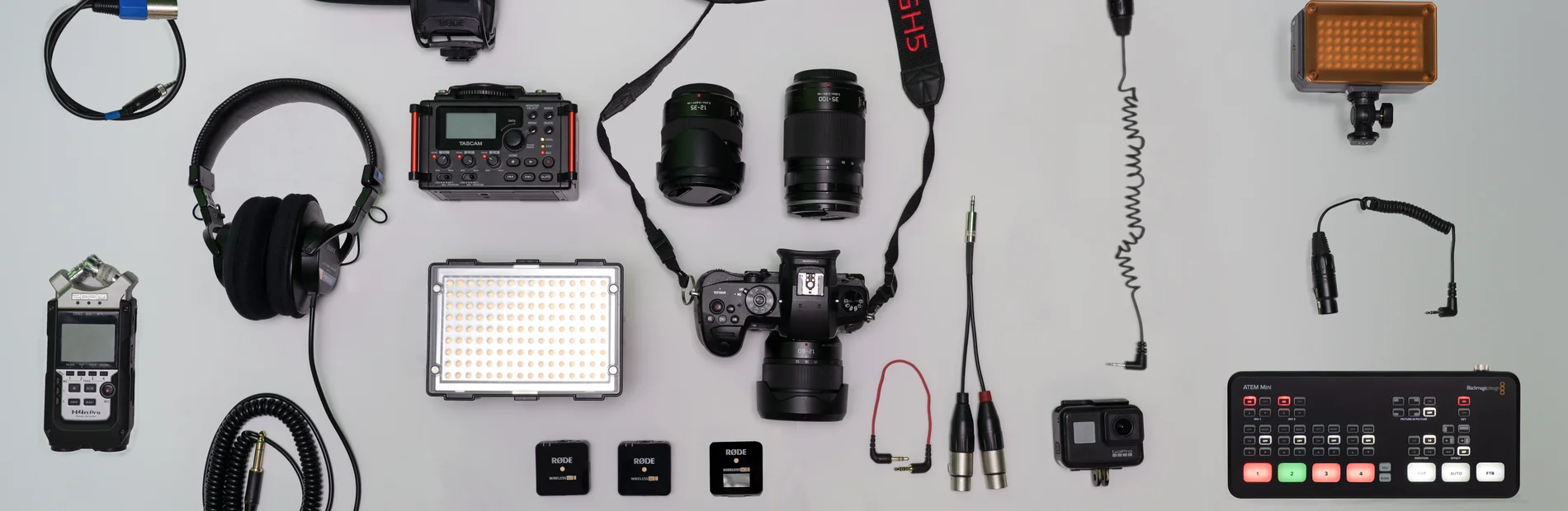 Verschiedene Kamera- und Audioausrüstungen, darunter Mikrofone, Objektive, Kopfhörer, LED-Leuchten und ein Mischpult, auf hellem Untergrund angeordnet.