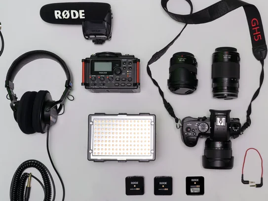 Verschiedene Kamera- und Audioausrüstungen, darunter Mikrofone, Objektive, Kopfhörer, LED-Leuchten und ein Mischpult, auf hellem Untergrund angeordnet.