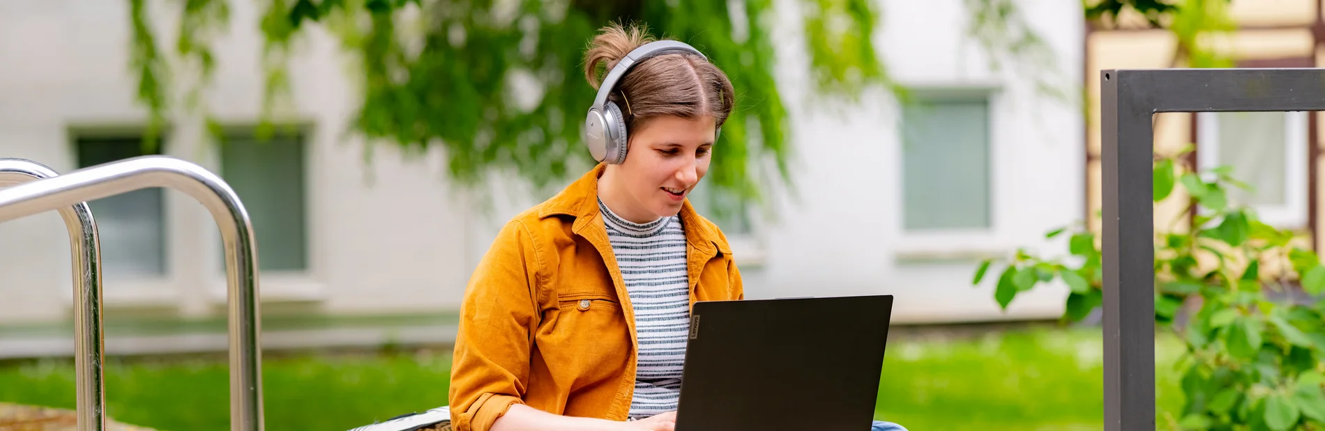 Junge Frau sitzt mit Laptop und Kopfhörern draußen