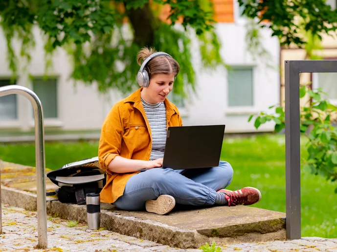 Junge Frau sitzt mit Laptop und Kopfhörern draußen