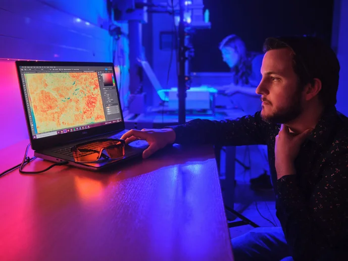 Digitale Rekonstruktion und Materialanalytik In pink- und violettfarbenem Licht bedient ein Mitarbeiter einen Computer, während im Hintergrund eine Mitarbeiterin eine Handschrift auf einer MSI-Anlage positioniert.