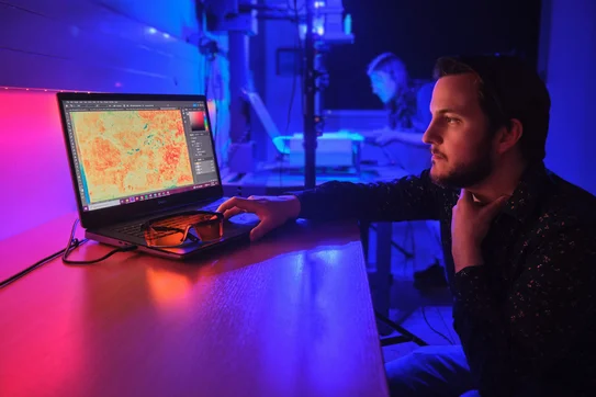 In pink- und violettfarbenem Licht bedient ein Mitarbeiter einen Computer, während im Hintergrund eine Mitarbeiterin eine Handschrift auf einer MSI-Anlage positioniert.