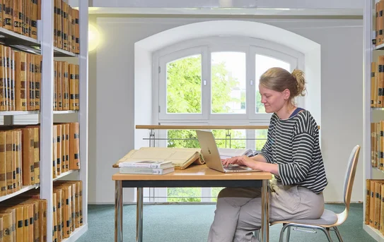 Nutzerin sitzt an einem Tisch zwischen Bücherregalen und tippt auf einem Laptop.