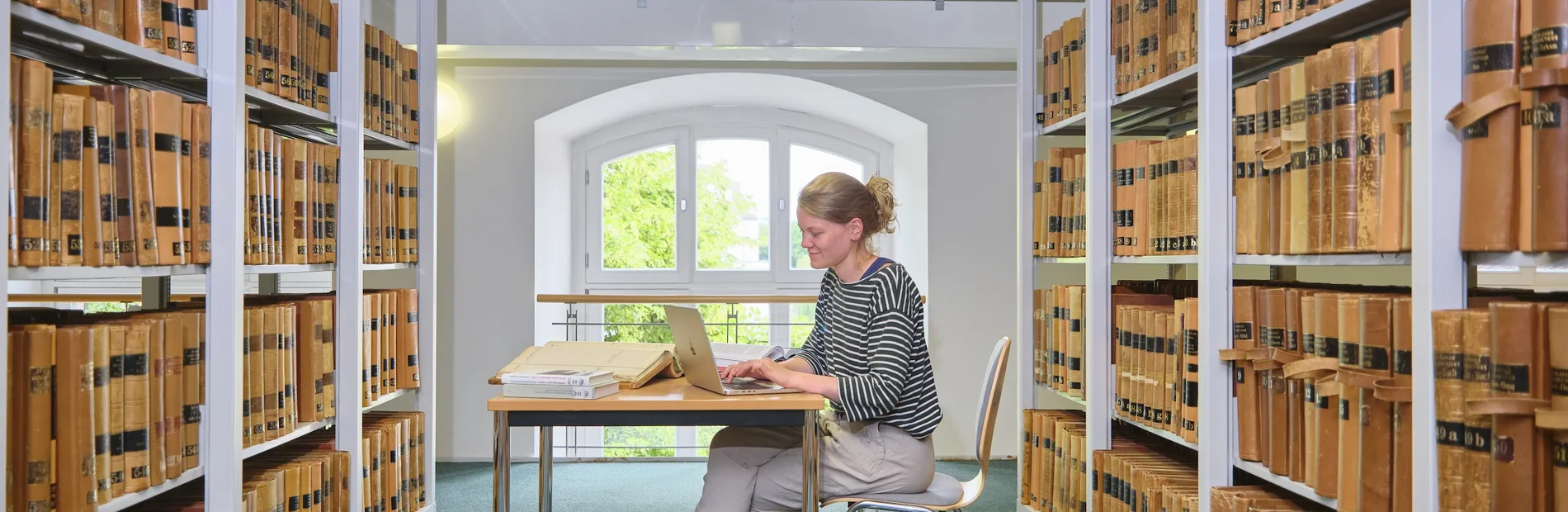 Besuch im Lesesaal Nutzerin sitzt an einem Tisch zwischen Bücherregalen und tippt auf einem Laptop.