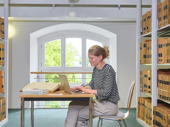 Besuch im Lesesaal Nutzerin sitzt an einem Tisch zwischen Bücherregalen und tippt auf einem Laptop.