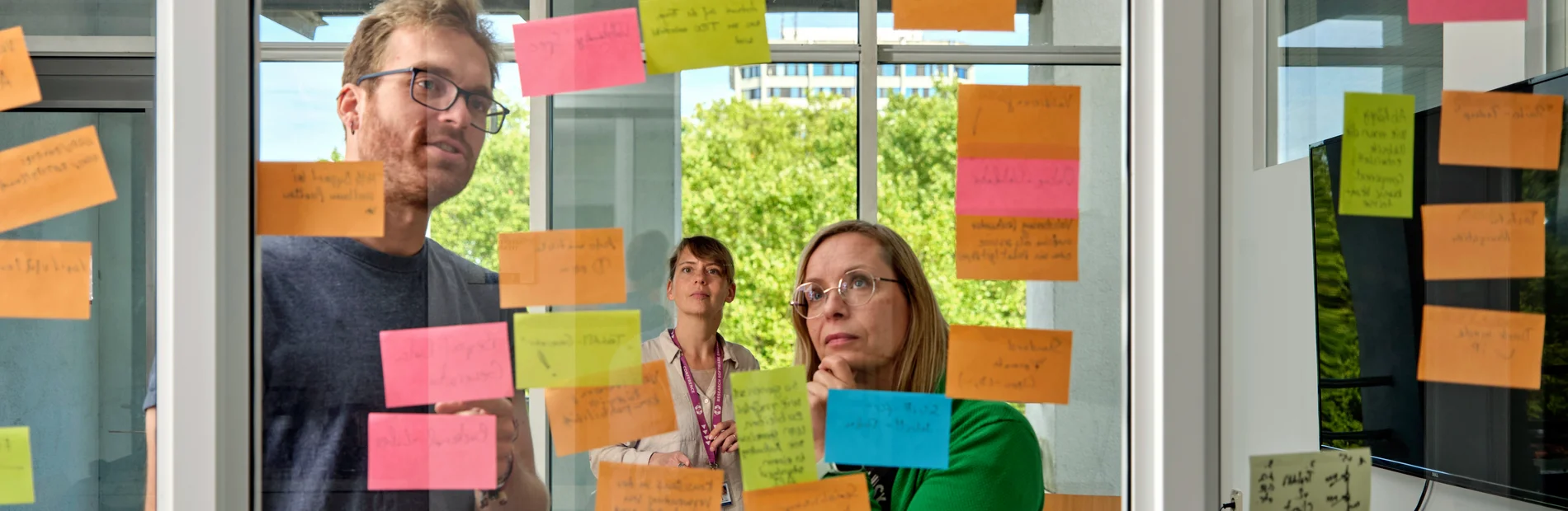 Forschungsservices_Header_20251118 Personen stehen vor einer Glaswand mit beschriebenen Notizzetteln und beraten sich.