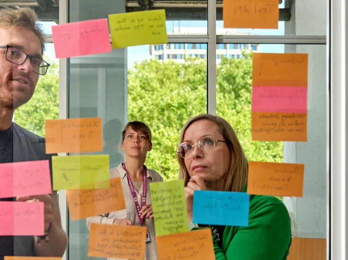 Personen stehen vor einer Glaswand mit beschriebenen Notizzetteln und beraten sich.
