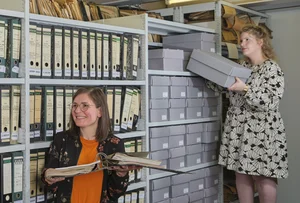 Drei Personen vor einem Regal mit Archivmaterial, eine Person nimmt eine Box heraus.