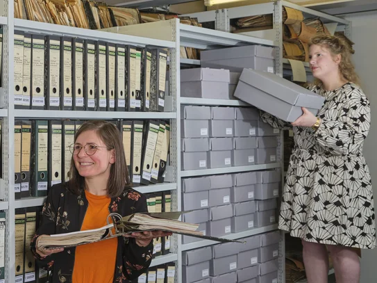 Drei Personen vor einem Regal mit Archivmaterial, eine Person nimmt eine Box heraus.