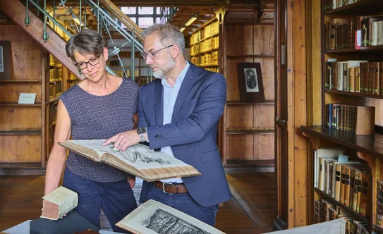 Zwei Personen im historischen Bibliothekssaal, eine zeigt der anderen ein altes Buch