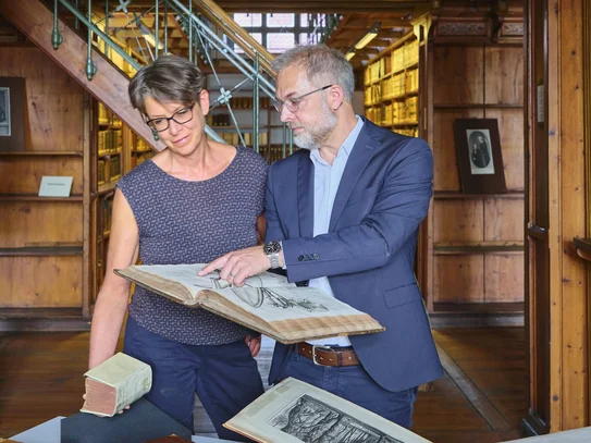Zwei Personen im historischen Bibliothekssaal, eine zeigt der anderen ein altes Buch