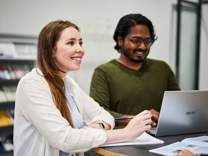 Zwei Personen stehen an einem Tisch, darauf steht ein Laptop. Eine Person hört aufmerksam zu