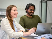 Zwei Personen stehen an einem Tisch, darauf steht ein Laptop. Eine Person hört aufmerksam zu